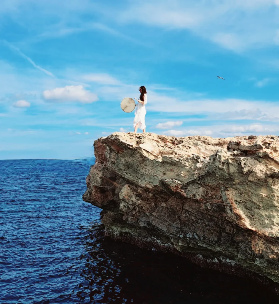 Frau steht auf einem Felsen am Meer und hält eine schamanische Trommel